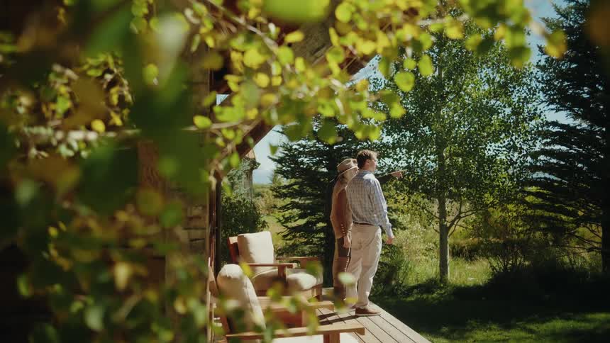 A couple stands on a patio on a sunny day two armchairs positioned behind them as they talk with a building surrounded by trees in the background