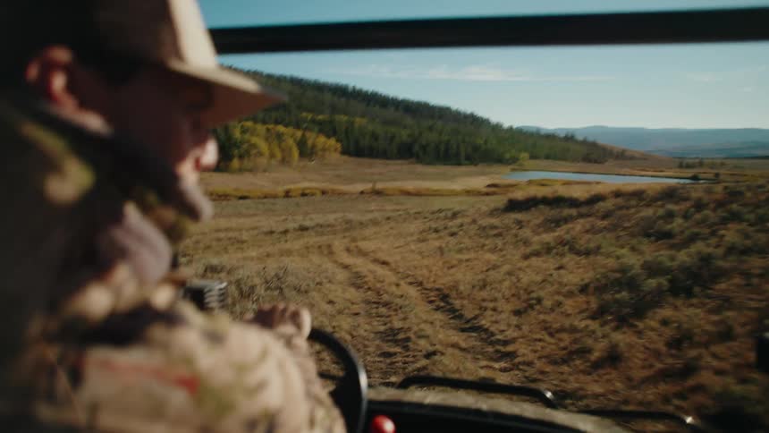 Adult young man wearing a baseball cap driving a vehicle through a rugged road near a pond with mountains on the background outdoors during day time