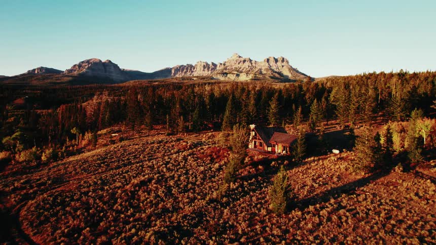 Isolated house in an open field surrounded by forest with a mountain range in the background under golden light