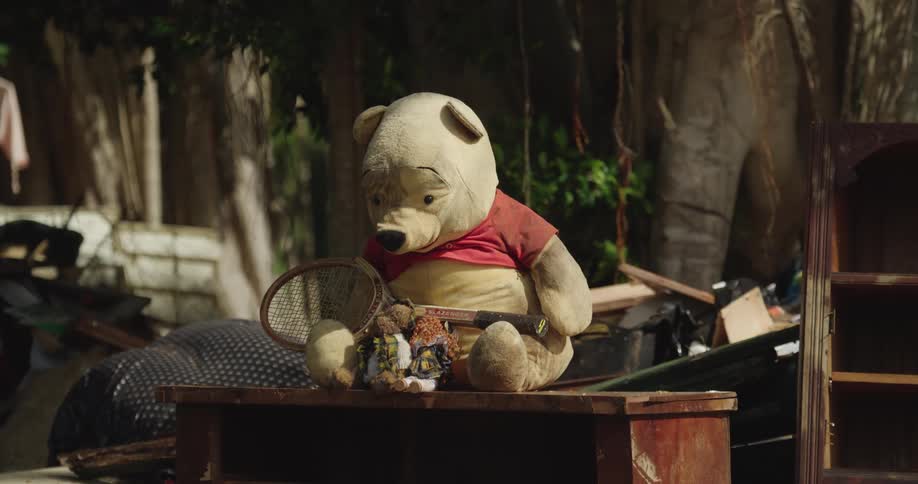 Teddy bear wearing a red shirt holds a racket while looking at small dolls on a table in an outdoor setting with surrounding objects
