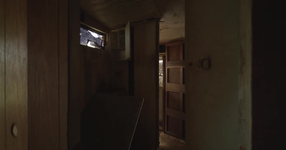 A view through the narrow and dark hallway of an abandoned house with peeling wallpaper and a broken window allowing dim light into the space