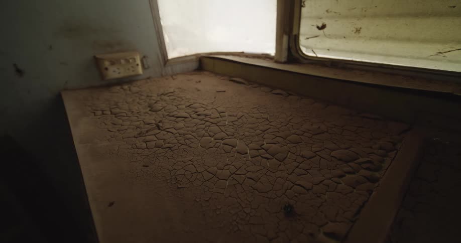 Close view of a thick layer of cracked dry mud covering a surface near a window in an abandoned room with soft natural light highlighting the texture