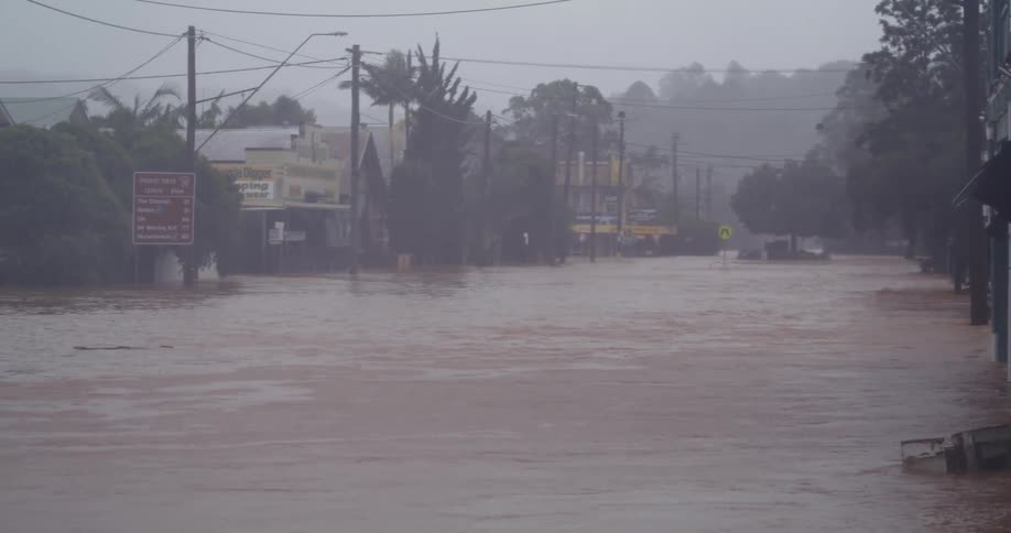 Brown murky floodwaters surge through a towns main street during a heavy downpour reaching the doorsteps of local businessesbuildings and power lines are visible along the flooded road partially obscured by the thick mist and falling rain