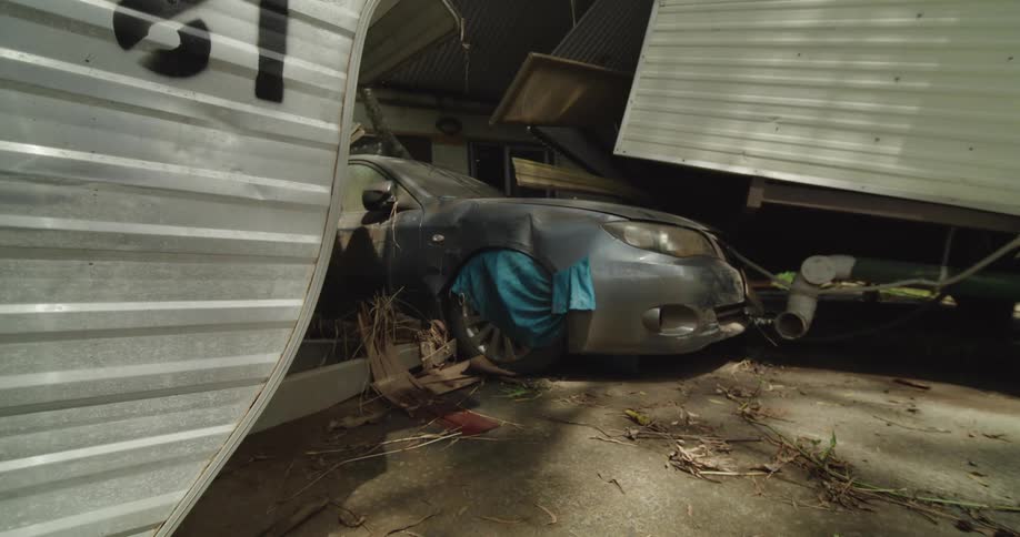 A garage with a scrap car parked inside its roof and hood covered with scattered rubble and debris