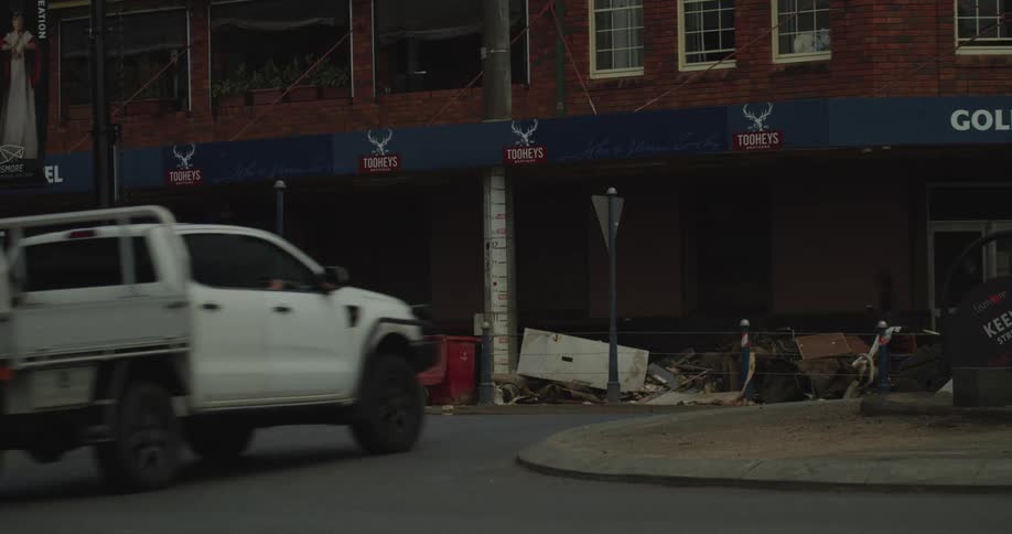 The video shows a street scene featuring a building with tooheys brewery signage above a pile of debris and discarded furniture on the sidewalk several vehicles including a white van a white sedan and a white pickup truck drive past the camera in the