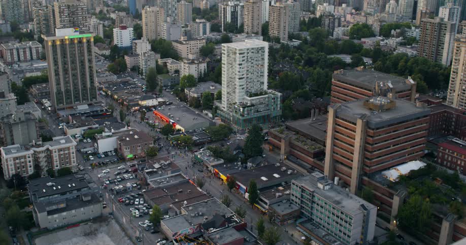 An aerial drone shot captures a bustling city neighborhood showing a dense mix of residential towers commercial storefronts with bright signs with streets flanked by parked vehicles and greenery