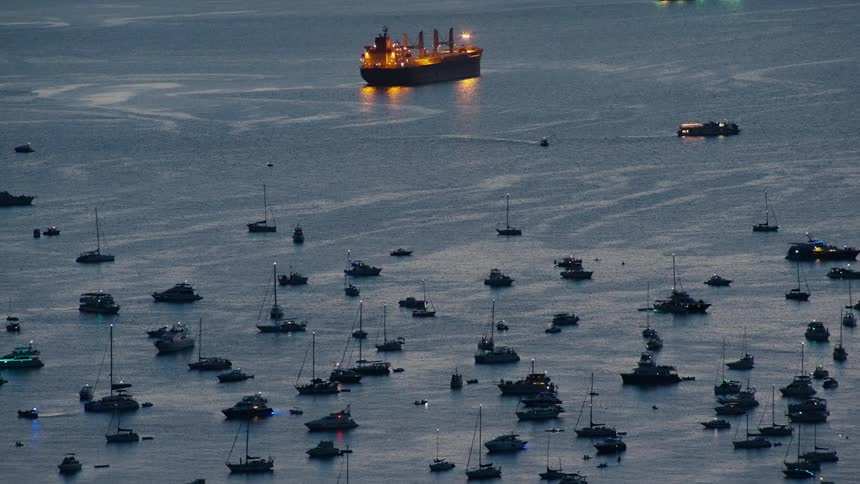 A distant view shows numerous small boats anchored in a quiet bay at dusk while a large cargo ship illuminated with yellow lights sits prominently in the background