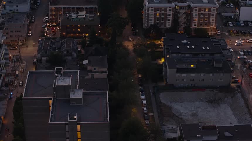 A nighttime city street with cars parked along the roadside as other vehicles pass by their headlights illuminating the urban scene