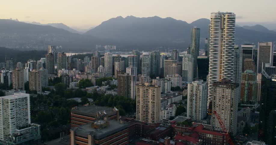 An aerial shot captures a dense cityscape with numerous high rise buildings and green spaces set against a backdrop of hazy mountains at dusk