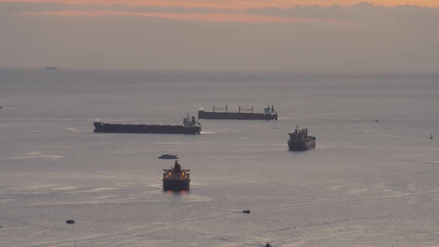 Wide view of large cargo ships anchored in the calm sea during a hazy sunset with small boats moving nearby and a soft golden light reflecting on the water