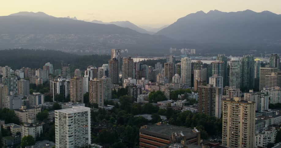 A wide aerial shot captures a dense downtown cityscape filled with buildings all set against the stunning backdrop of hazy mountains during a beautiful soft sunset