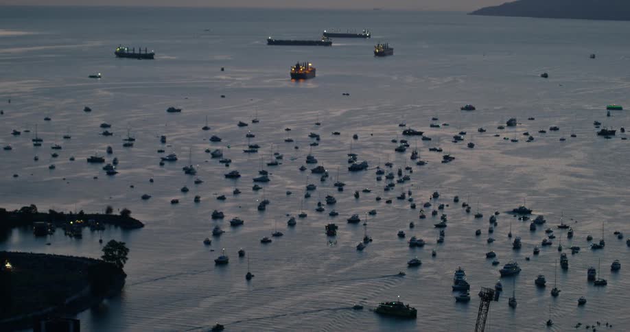 Dozens of small boats and several large cargo ships sit anchored in a harbor during the blue hour as smaller vessels navigate through the crowded water near a dark forested shoreline