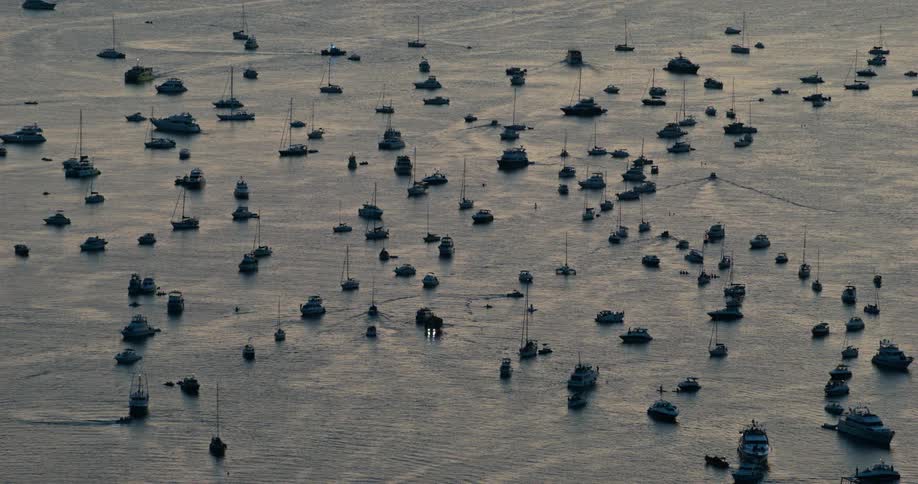 An aerial view shows a dense cluster of sailboats and motorboats anchored in a calm body of water during twilight the scene captures a tranquil atmosphere with small ripples on the waters surface as boats of various sizes are scattered across the bay