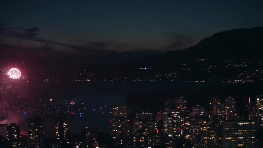 Aerial panning view of fireworks exploding over a city harbor with boats and illuminated skyscrapers