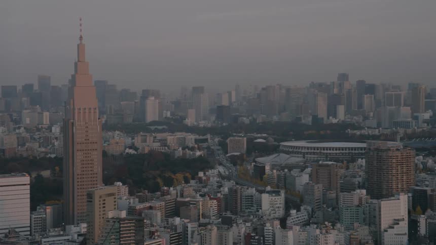 An aerial view captures the vast dense cityscape of tokyo during the hazy twilight hoursthe camera pans across a landscape of endless buildings dominated by a tall spiretopped skyscraper and a large stadium nestled near a forested park