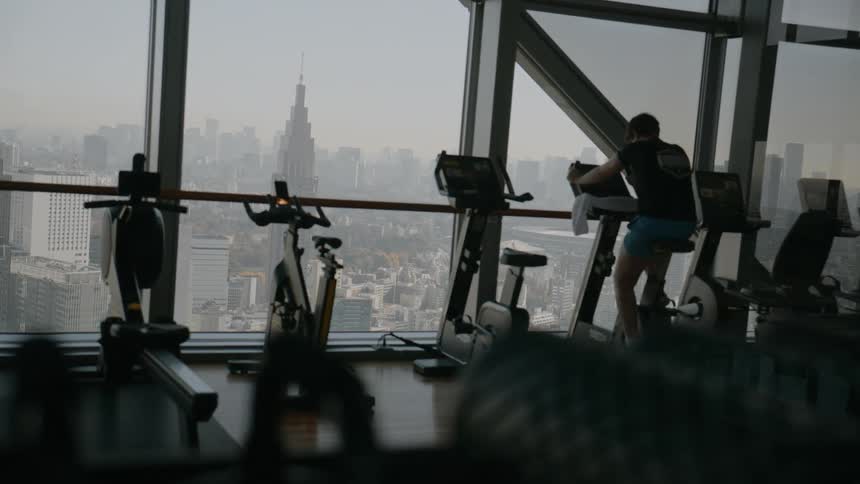 Young adult man riding a stationary bike at a gym in front of a glass window showing a metropolitan area indoors during day time