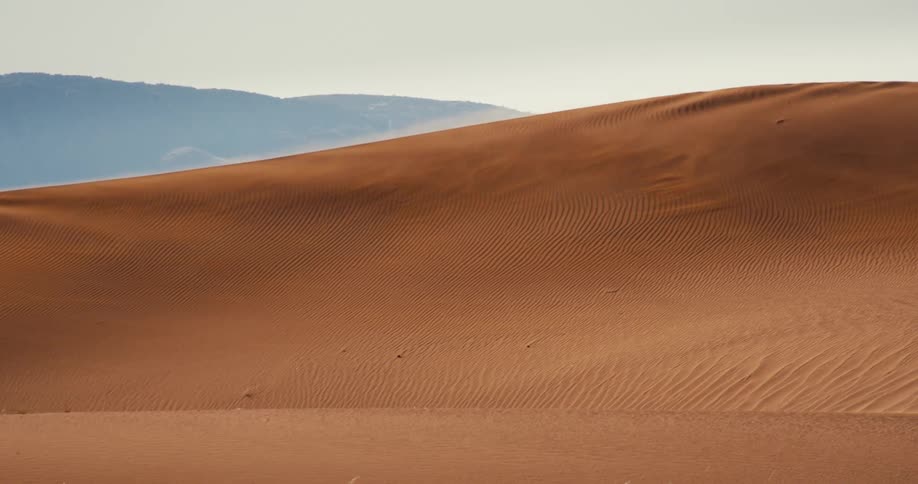 Sand blows across a desert dune with mountains in the distance