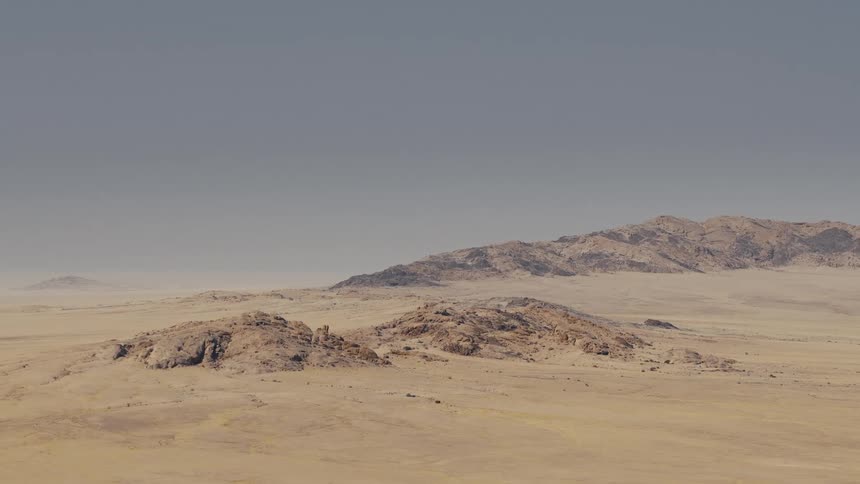 A distant view shows a vast arid desert landscape under a hazy gray sky with several low rocky mountain ridges scattered across the sandy terrain