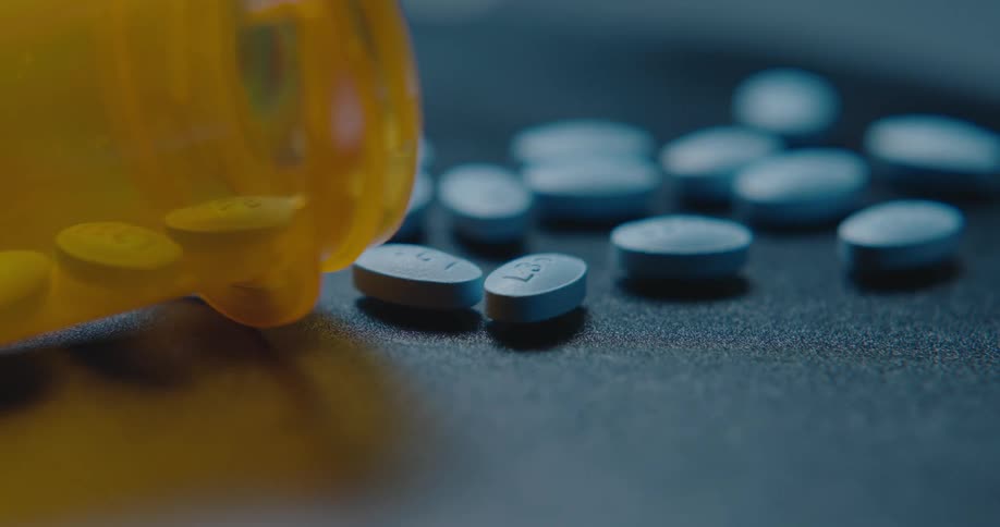 A closeup view shows an orange pill bottle lying on its side next to several blue ovalshaped pills scattered across a dark textured surface