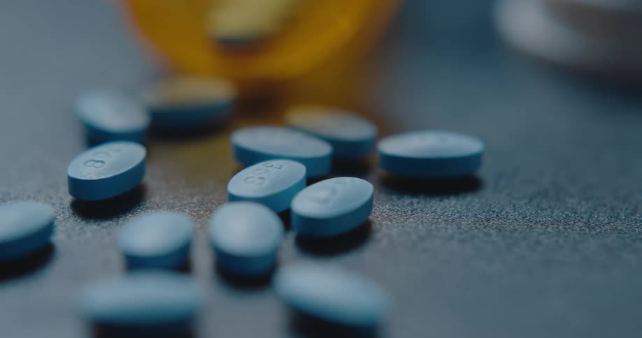 A close view shows a textured dark surface with a white bottle cap in the foreground and blurred blue pills near an orange container in the background