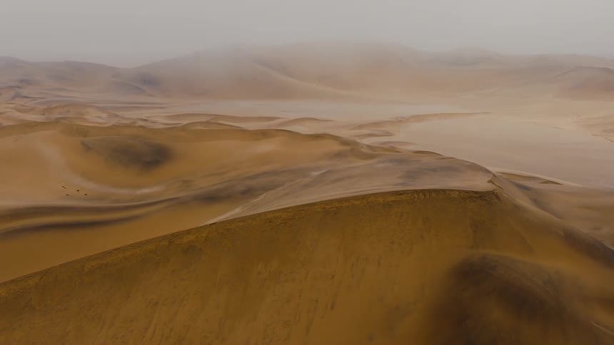 An aerial drone shot pans across a vast desert revealing undulating sand dunes and misty mountains under a hazy sky