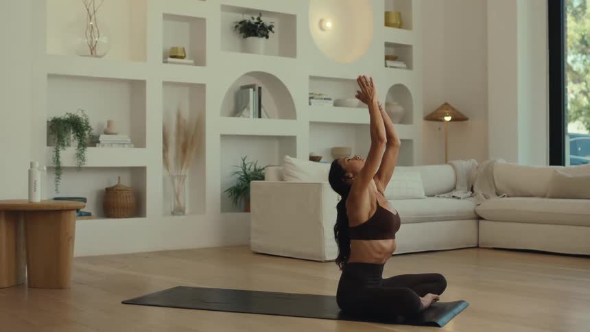 A woman sits on a black mat in a bright living room raising her arms overhead with palms together before lowering them to her chest in a prayer position to meditate