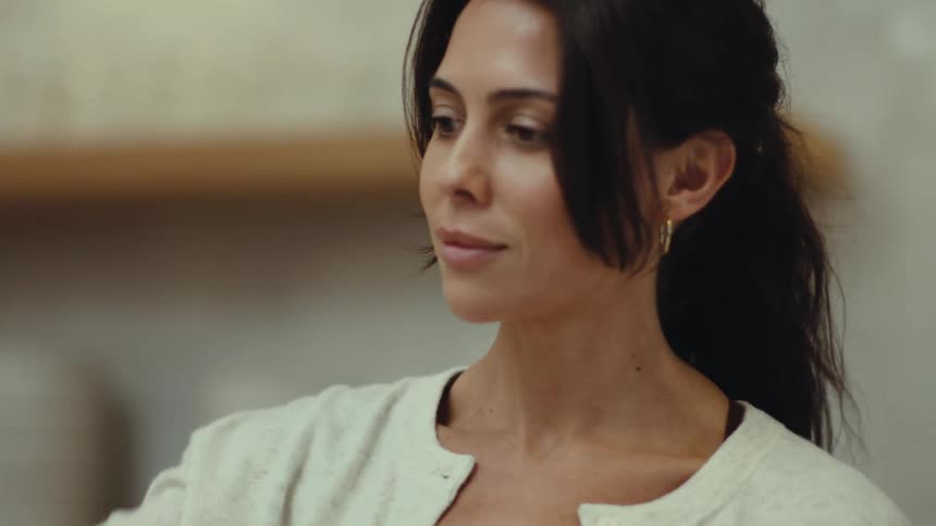 A woman with dark hair tied back wears a white cardigan and gold earrings while standing in an indoor setting looking down with a neutral expression as she moves slightly