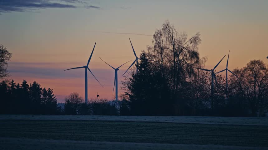 Wind turbines on a wind farm surrounded by trees with a natural landscape on the background outdoors during dusk or dawn