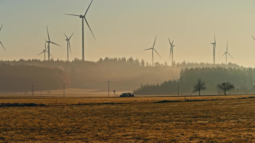 Cars drive along a road crossing a field during golden hour with a dark forest line and large wind turbines silhouetted against a hazy sky in the background