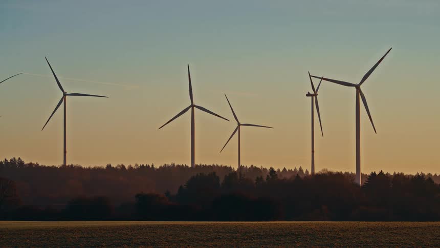 Tall wind turbines stand atop a forested ridge during a golden dusk