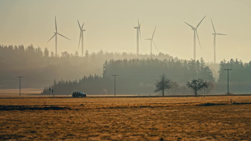 A car drives down a rural road through a hazy golden landscape with a forest and a wind farm visible in the background