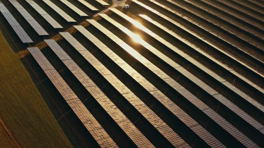 An aerial drone shot glides over rows of solar panels at a large power station during sunset capturing the golden sunlight reflecting off the glass surfaces as the camera moves across the vast energy field
