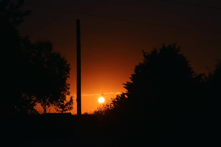 A stationary wide shot captures a bright orange sun descending toward the horizon during dusk casting dark silhouettes of trees a utility pole and distant power lines against a deep gradient sky