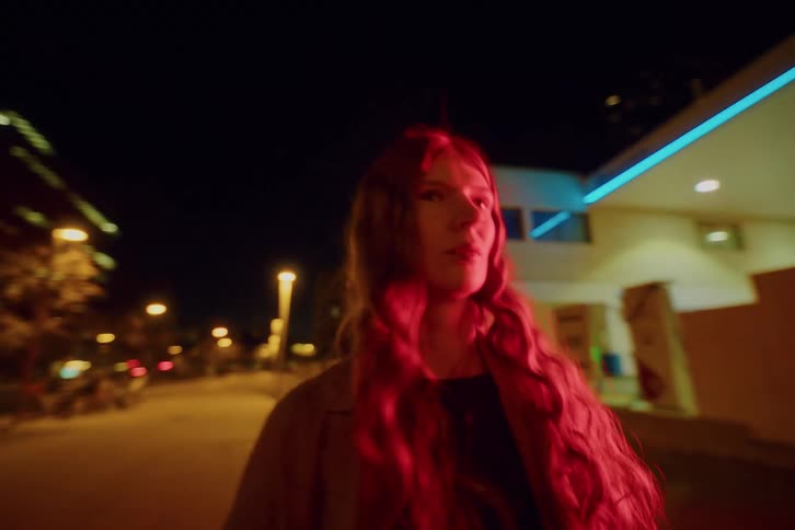 A young woman standing in the middle of the street at night illuminated by the bright glow of a nearby sign reflecting on her