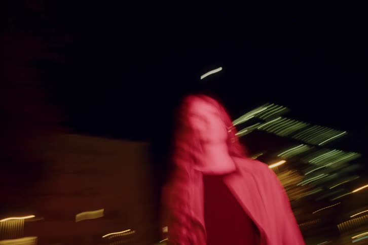 A young woman walks along a nighttime street wearing a jacket illuminated by the warm glow of building lights streaming across the pavement