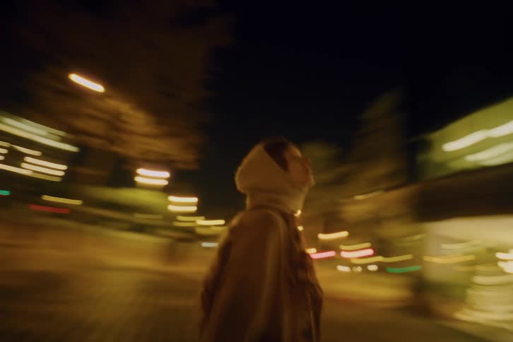 Female person wearing a scarf over her head walking on a street next to a park on an urban area outdoors during night time