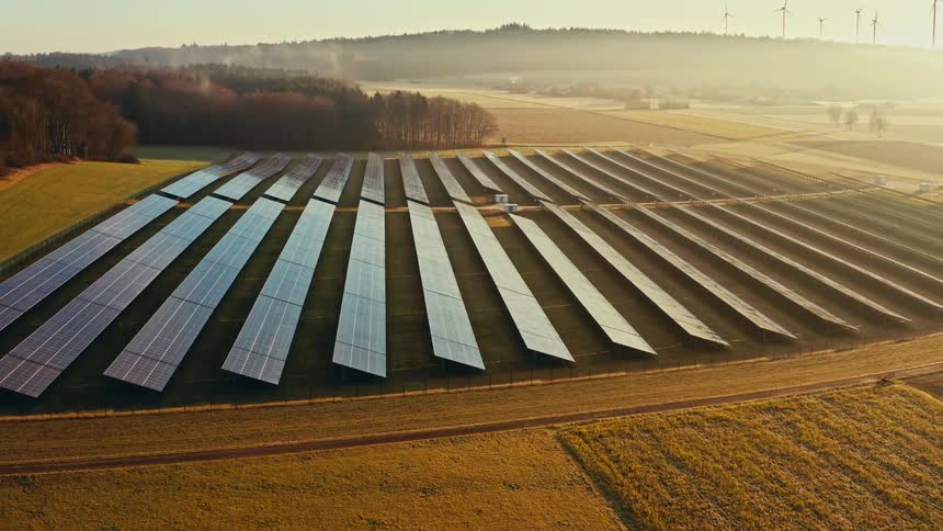 An aerial drone shot pans across a large solar farm situated in a rural valley at sunset showing rows of panels reflecting the low sun while wind turbines spin on a hazy ridge in the distant background