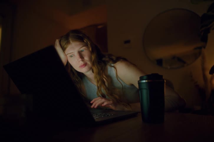 In a dimly lit room at night a tiredlooking woman with long hair leans her head on one hand while staring intently at a laptop screen a reusable coffee cup beside her suggesting latenight work or study