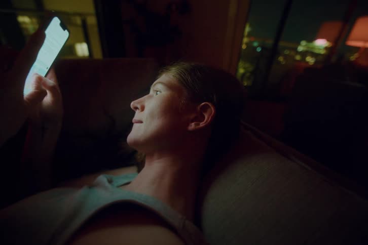 Person lying indoors at night looks at a glowing phone screen with city lights in the background