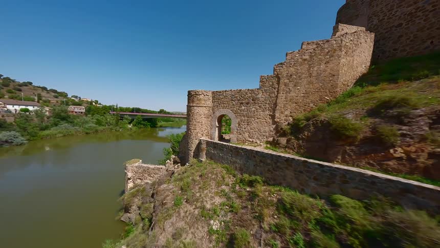 A sunlit path leads toward a distant castle with a calm river running alongside it as the bright day highlights the landscapes warm