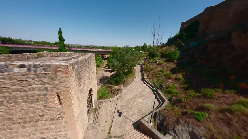 An fpv drone captures a dynamic aerial view as it glides over a wide greenish river toward a historic stone fortress the camera banks sharply revealing a modern bridge in the background and ancient defensive walls built along the rocky shoreline