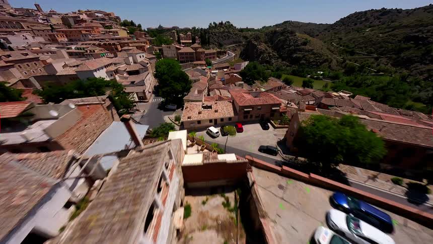 An aerial fast view flies over the brown tiled roofs of a historic city and descends towards a winding road with cars near a river under a clear blue sky