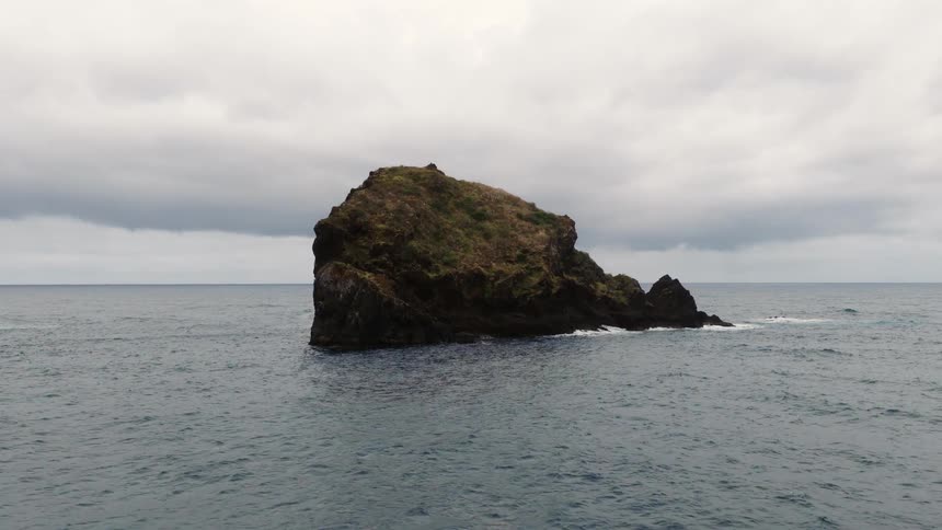 A large isolated sea stack covered in sparse greenery as it sits amidst choppy dark ocean water under a heavy overcast sky