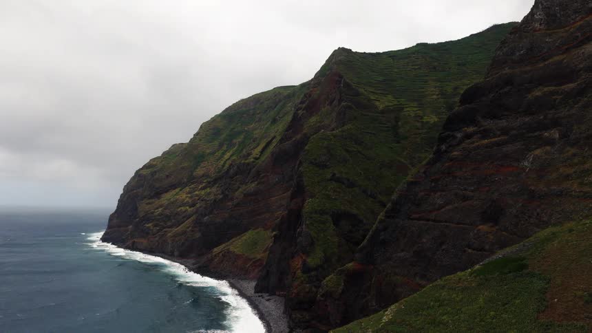An aerial shot shows massive greencovered cliffs meeting the dark ocean with waves crashing against a narrow pebble beach at the base of the steep mountains under a heavy overcast sky