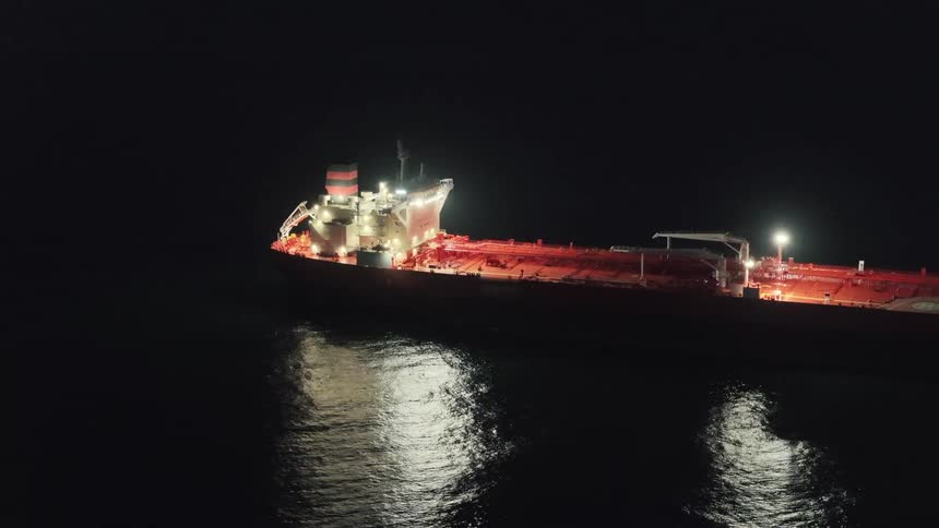 The video features a massive oil tanker floating on calm dark waters during the nightthe ships superstructure is brightly illuminated casting a shimmering golden reflection across the surface of the sea