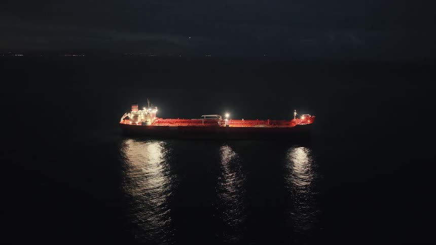 A large industrial tanker ship illuminated by bright deck lights sits anchored on dark calm water at night under a pitch black sky