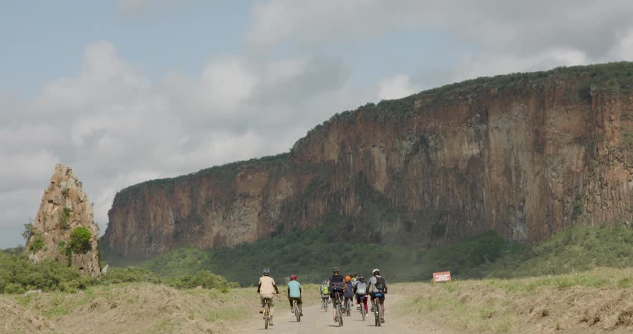 A group of cyclists rides away from the camera down a dirt path toward a massive flattopped cliff under a cloudy sky