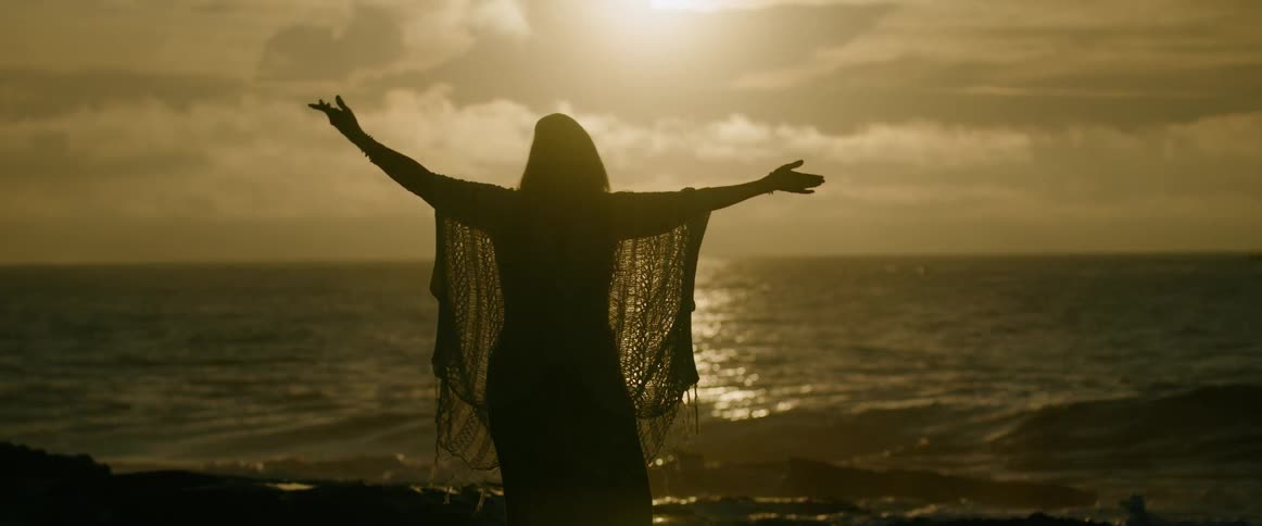 A young woman stands on the beach at sunset lifting her arms toward the sky as waves roll in and break along the glowing shoreline