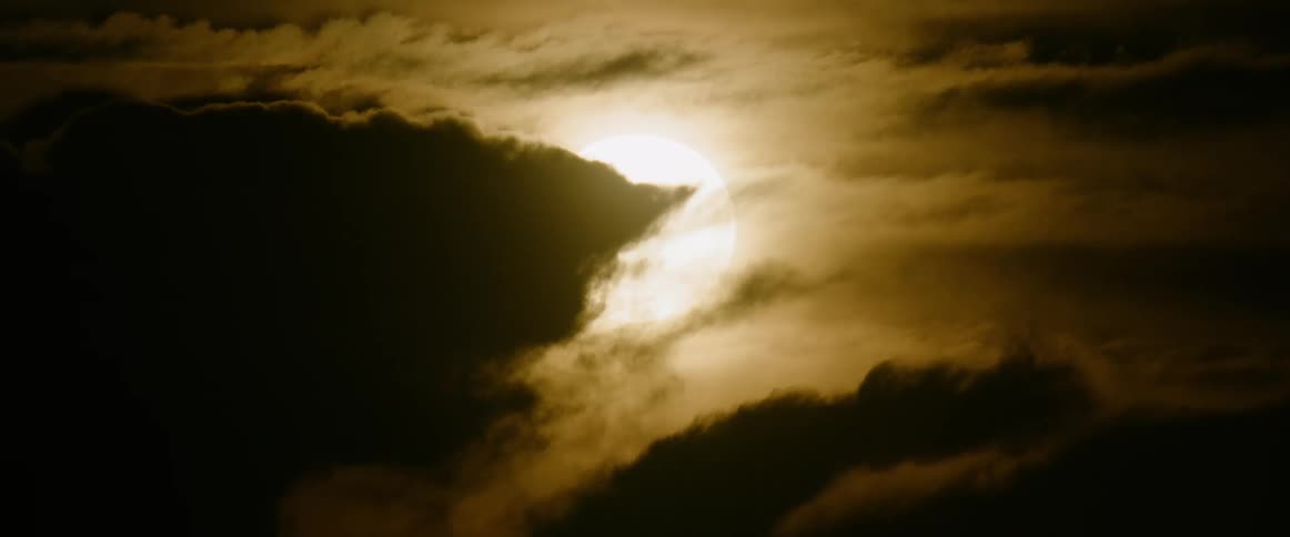 A closeup view shows a large bright sun partially obscured by heavy dark golden clouds that drift slowly across the frame under a hazy sky
