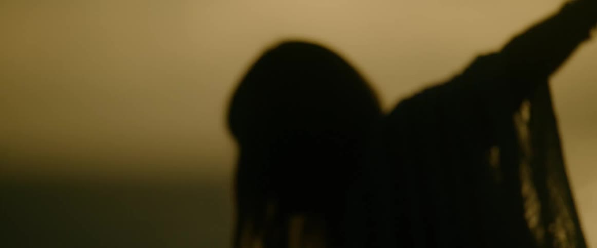 A woman is seen in silhouette dancing against a golden twilight sky gracefully moving her head and raising her arms upward while wearing a lace shawl that catches the fading light